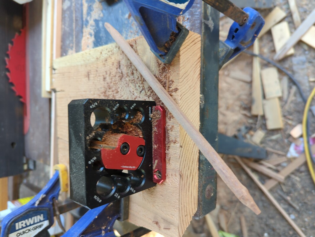 small dowel cutting jig clamped to a sawdust covered work table. Aside the jig is freshly cut 3/8 inch diameter dowel of Redwood