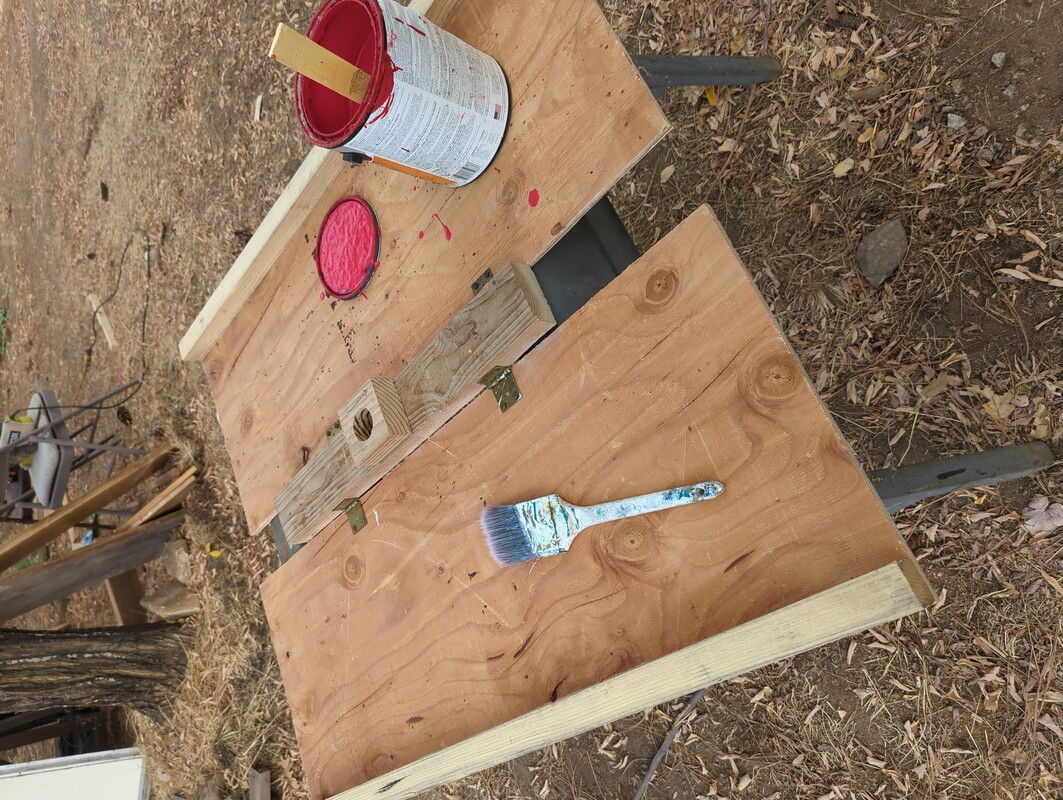 flat plywood panels attached to both sides of a short length of 2x4. There is a gallon bucket of paint, and a paint brush, resting on the panels.