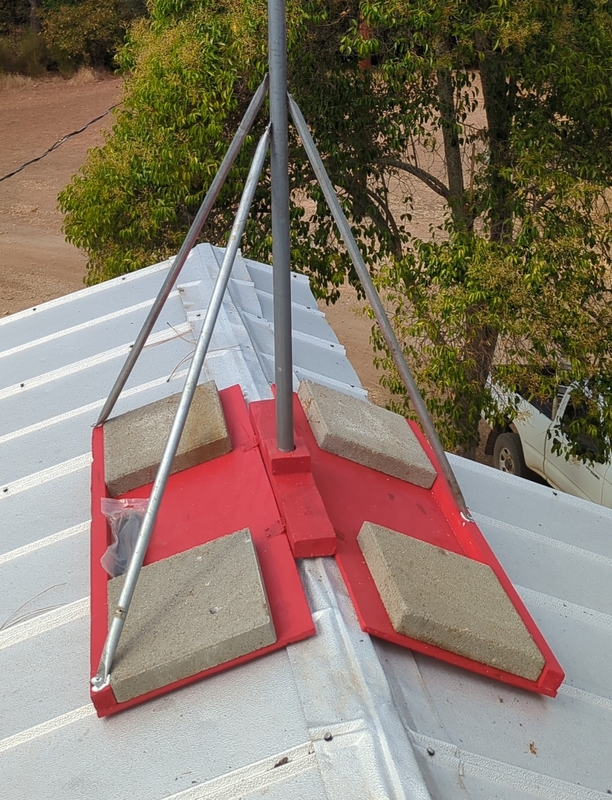 painted base in position on roof ridge. A pole is in the mount socket, and 3 metal supports extend from the middle of the pole to the ends of the base. the base is held down by 4 concrete pavers