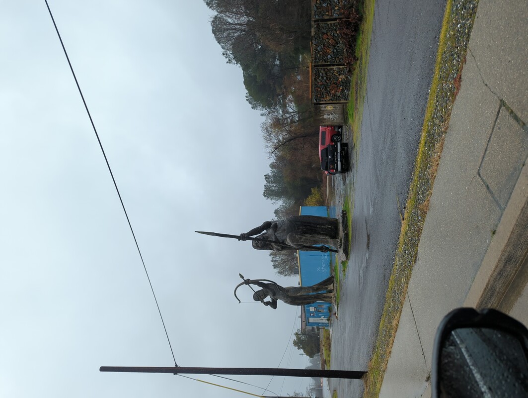 two 20ft tall concrete statues of warrior women next to a road. One is standing using a bow and arrow, the other is kneeling and holding a spear. There is a warehouse behind the statues. The sky is overcast and everything is wet.